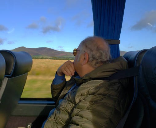 Κάθεται ήσυχα, φοράει γυαλιά ηλίου, ακούει τους γύρω χωρίς να μιλά.